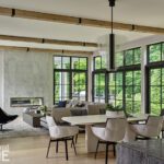 Open-concept great room with modern dining and living areas, exposed wood ceiling beams, and floor-to-ceiling black-framed windows overlooking greenery.