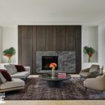 Contemporary living room with dark wood-paneled fireplace, neutral furnishings, and large windows creating a seamless indoor-outdoor connection.