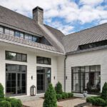 Private courtyard patio of a modern Greenwich home featuring white shingle siding, large black-framed windows, and manicured formal plantings.