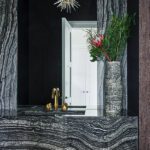 Stylish powder room with dramatic black and gray veined stone vanity, brass fixtures, textured wallpaper, and sculptural pendant light.
