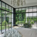 Serene primary bedroom with floor-to-ceiling glass walls offering panoramic views of lush greenery and patio, styled with a modern chandelier and cozy neutral bedding.