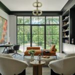 Contemporary home study with modern furnishings, built-in shelves, large abstract art, and full-height windows overlooking a wooded landscape.