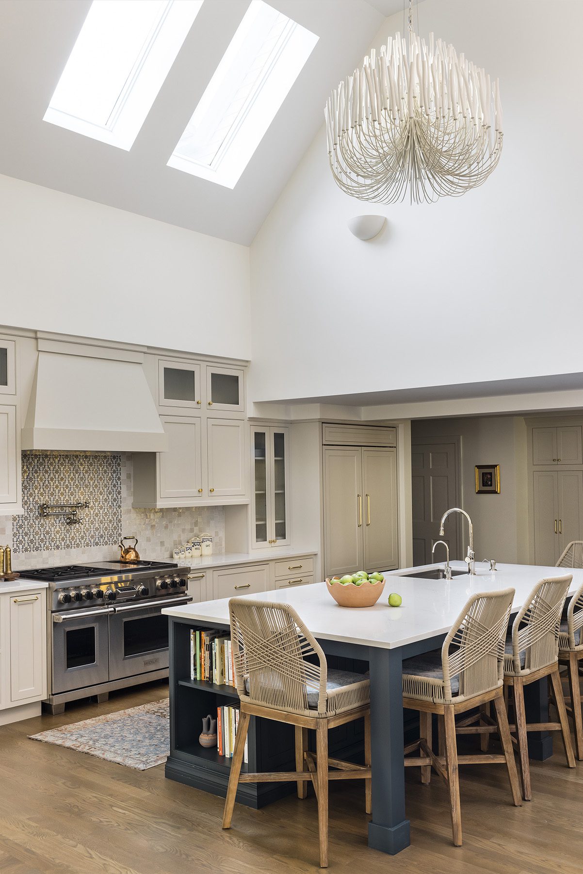 Bright modern kitchen with skylights, white cabinetry, and large island featuring woven bar stools and a unique chandelier