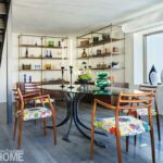Glass-top dining table with colorful upholstered chairs surrounded by brass and wood shelving filled with art glass and vintage pottery.