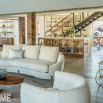 Open-concept interior blending living and dining areas with vintage glass decor, soft boucle seating, exposed wood beams, and custom shelving.