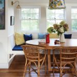 Cozy built-in breakfast nook with a round wood table, wicker chairs, floral pendant light, and colorful throw pillows beneath sunlit windows.