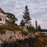 Shingle-style coastal home perched on a rocky Maine shoreline with pine trees and sweeping ocean views at sunset.