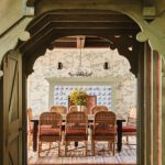Charming dining room with wallpapered walls, wicker chairs, rustic wood table, and a blue-and-white tiled fireplace framed by green wood archways.