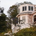 Classic shingle-style cottage with an octagonal sunroom and white trim nestled in lush greenery overlooking the coast.