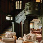 Warm and rustic great room featuring a dramatic green-painted staircase, cathedral ceilings, and upholstered armchairs with floral pillows.
