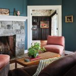 Cozy living room with deep teal walls, a rustic stone fireplace, and warm-toned seating accented by striped and floral pillows.