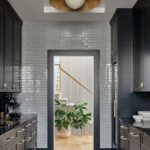 Sleek butler’s pantry with glossy gray subway tile walls, black cabinetry, brass hardware, and a natural wood floor in a luxury coastal home.