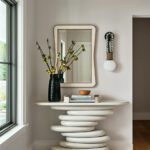 Stylish foyer with sculptural console table, decorative mirror, black vase with flowers, and modern wall sconce in a light-filled entryway.