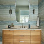 Serene guest bathroom with wood vanity, sculptural mirror, ocean-inspired wallpaper, and modern wall sconces in a luxury coastal residence.