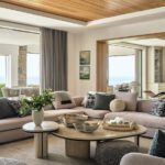 Elegant living room with plush sectional sofa, round nested coffee tables, wood ceiling, and ocean views in a contemporary New England home.