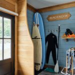 Beach gear storage room with surfboards, wetsuits, paddles, and snowshoes neatly organized in a wood and blue-paneled nook labeled “Beach Nook.”