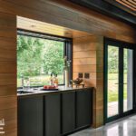 Contemporary indoor bar area with wood-paneled walls, black cabinetry, and a passthrough window offering scenic views of lush greenery.