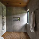 Minimalist bathroom with gray tile walls, teak slatted floor, rainfall showerhead, and a window overlooking the natural surroundings.