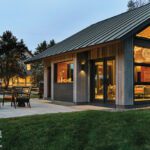 Twilight view of illuminated party barn with large windows, cozy interior glow, and an outdoor seating area with a lit modern fire pit.