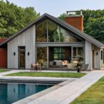 Modern party barn with pitched roof and expansive glass facade overlooking a sleek in-ground pool surrounded by stone paving and manicured lawn.