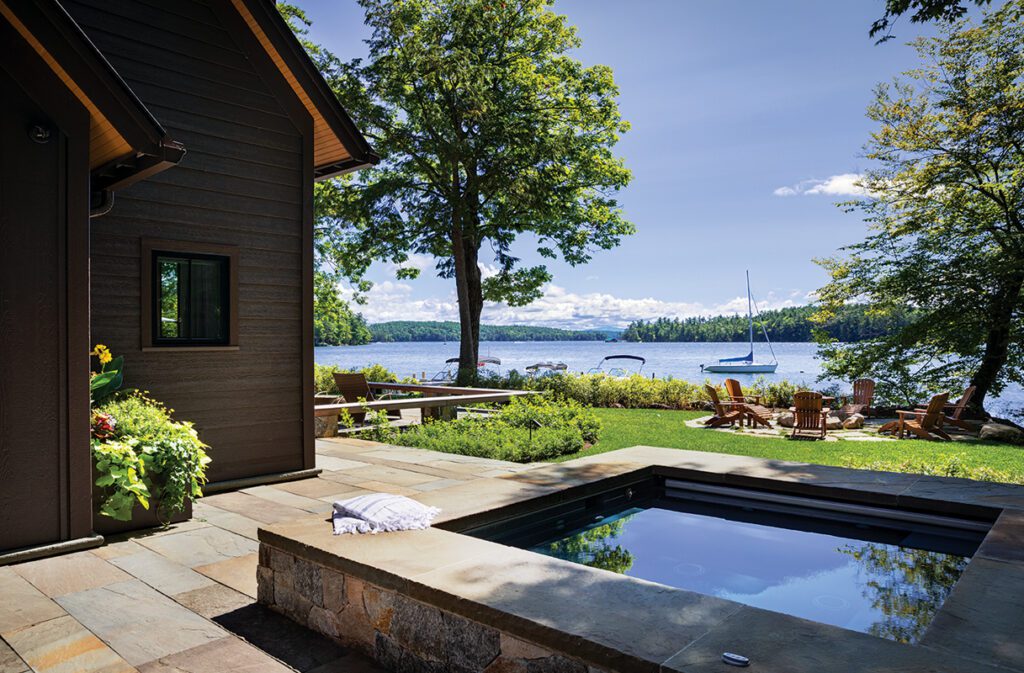 Elegant plunge pool with stone surround overlooking a scenic lake, surrounded by lawn, lounge chairs, and mature trees.