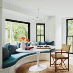 Bright bay window breakfast nook with a curved bench seat, blue cushions, and a white tulip table surrounded by nature views.