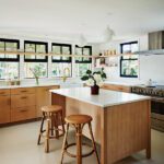 Contemporary kitchen with light wood cabinetry, white countertops, a central island with stools, and black-trimmed windows.