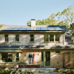 Cedar-shingled Cape Cod home with solar panels, green trim, and a welcoming entry flanked by beach towels and a vintage bike.