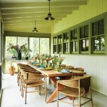 Screened-in porch with a long wooden dining table, woven chairs, and green-trimmed windows, set for an inviting alfresco gathering.
