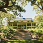 Rear view of a shingle-style home with a wide staircase leading to a screened porch, surrounded by lush landscaping and mature trees.