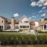Shingle-style Cape Cod home with stone chimneys, expansive windows, and a beautifully landscaped backyard with patio seating.
