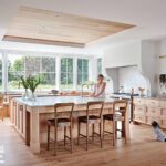 Bright and airy kitchen featuring a large island, wood cabinetry, and floor-to-ceiling windows overlooking a lush garden.