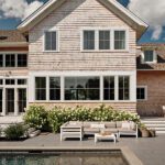 Elegant poolside patio with sectional seating, lush landscaping, and shingled Cape Cod architecture in the background.