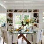Light-filled dining area with a round glass table, slipcovered chairs, and white built-in shelving showcasing curated vintage decor.