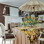 Eclectic dining room featuring a floral tablecloth, vintage cane chairs, and a dramatic indoor parasol centerpiece above the table.