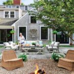 Cozy backyard firepit area surrounded by wicker seating and a shingle-style home, with a woman and two dogs near the outdoor lounge.