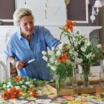 Woman arranging vibrant orange and white flowers on a colorful tablecloth beneath a decorative indoor parasol in a charming dining room.