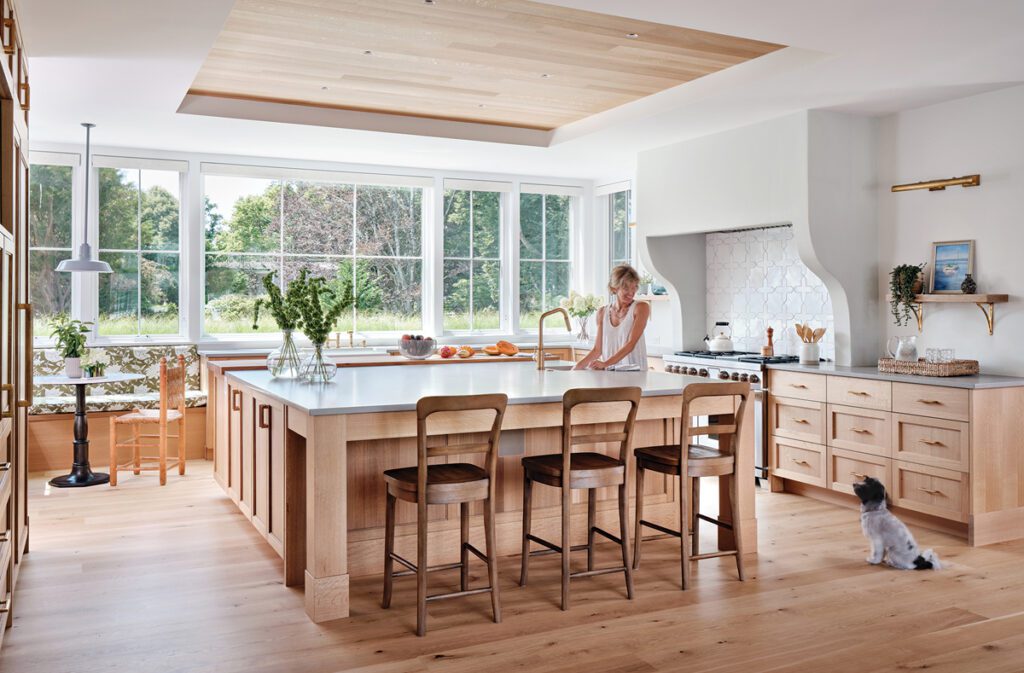 Bright and airy kitchen featuring a large island, wood cabinetry, and floor-to-ceiling windows overlooking a lush garden.