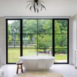 Freestanding white tub in a serene modern bathroom with large black-framed windows and a view of the green backyard.
