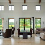 Airy modern living room with beamed ceiling, stylish seating, and black-framed windows opening to green views.