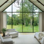 Spacious primary bedroom with vaulted ceiling, exposed wood beams, and dramatic floor-to-ceiling windows showcasing lush green views.