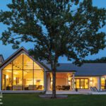Rear view of a modern farmhouse at twilight with expansive windows, warm interior lighting, and a mature tree in the foreground.