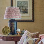 Close-up of a traditional side table with a black and gold chinoiserie lamp, antique artwork, and floral pillows on a pink sofa.