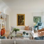 Airy and modern living room with white and orange seating, glass coffee table, and a large landscape painting over the sofa.