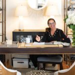 Living Swell shop owner at her desk surrounded by fabric samples, fresh flowers, and shelving filled with baskets and pillows in a chic office nook.