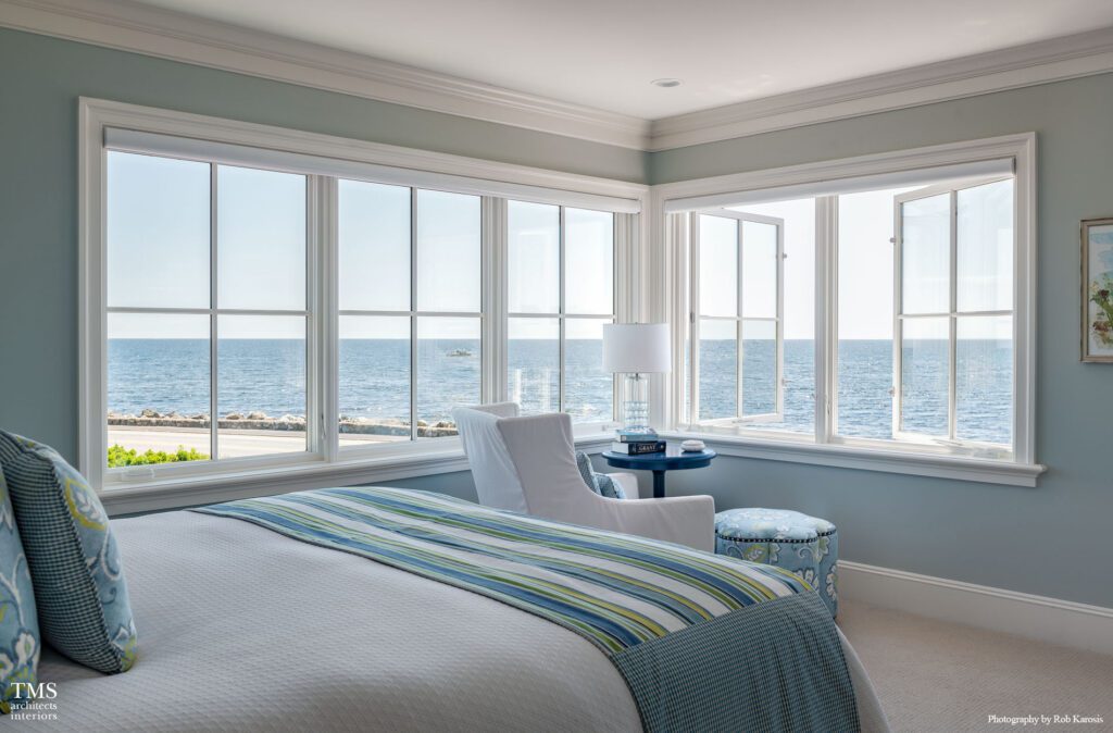 Coastal bedroom with panoramic ocean views, featuring soft blue walls, striped bedding, and a cozy reading nook by expansive corner windows.