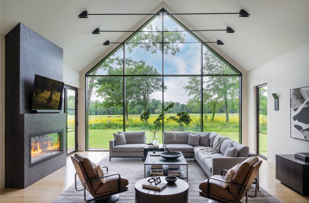 Bright, modern living room with vaulted ceiling, sleek fireplace, and floor-to-ceiling windows revealing a scenic meadow.