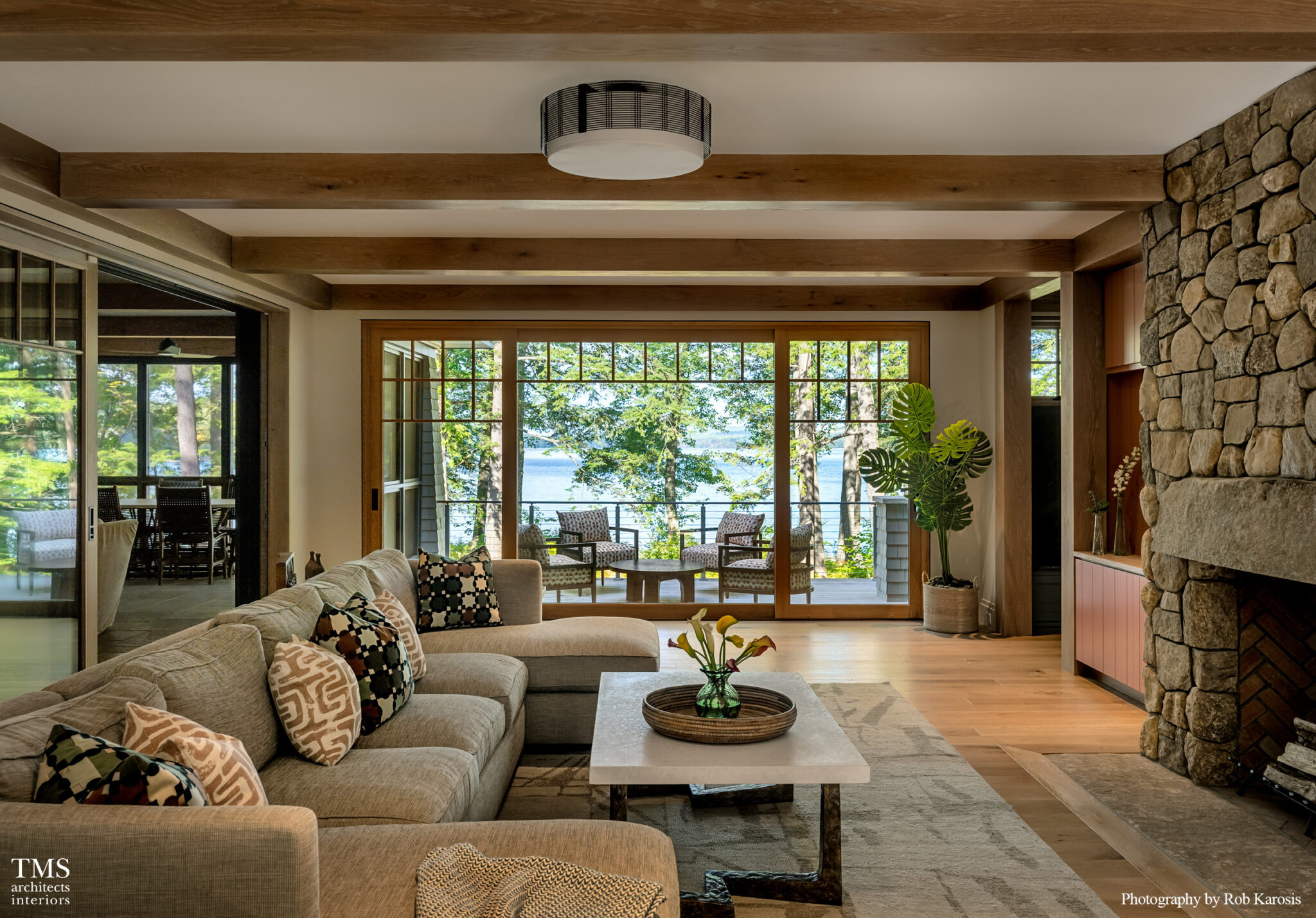 Warm living room with rustic stone fireplace, exposed wood beams, and sliding glass doors leading to a forested lakefront patio.