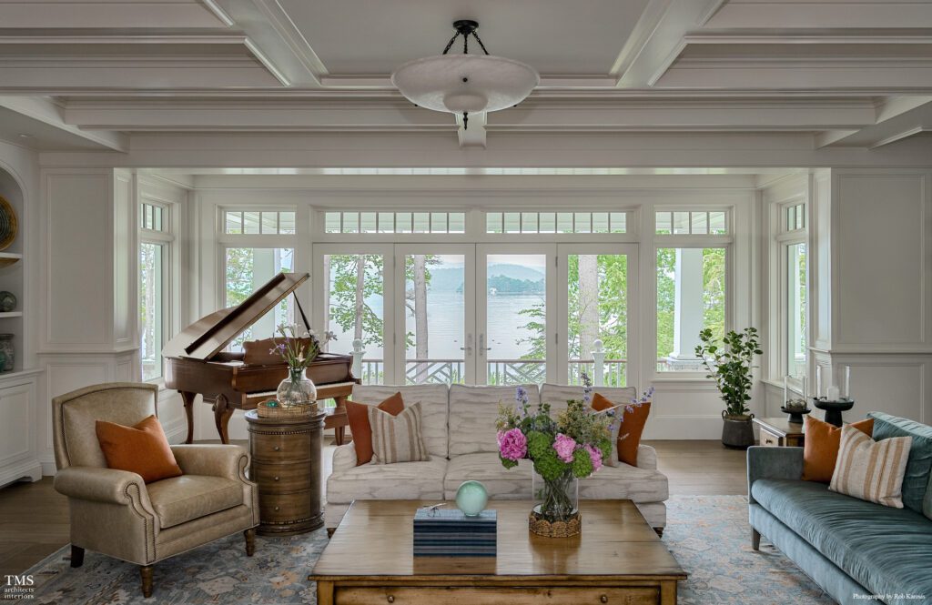 Elegant lakeside living room with coffered ceiling, grand piano, and large windows framing a scenic waterfront view surrounded by trees.