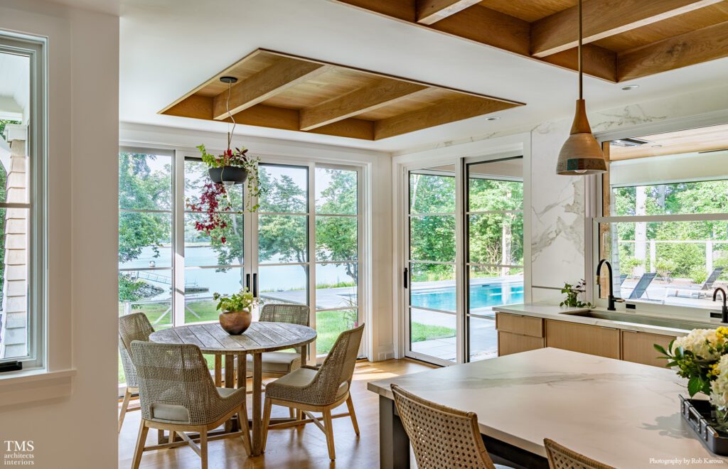 Modern breakfast nook with rattan chairs and round wood table overlooking a serene backyard pool and water view through floor-to-ceiling windows.